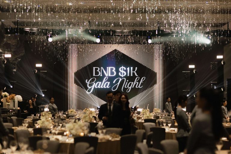 Wide shot of the main ballroom setup for BNB 1K Gala Night hosted by The Brooker Group PCL, featuring a illuminated stage screen and tables set for dinner.