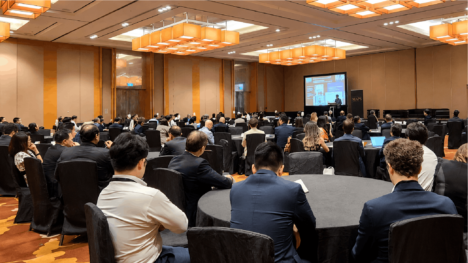 Wide shot of a large audience seated at round tables listening to a presenter on stage at the MAPS by BluFin conference.
