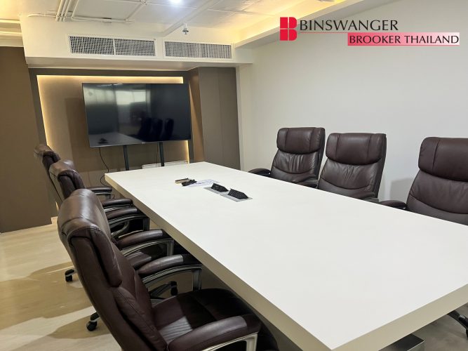 A modern conference room featuring a long white meeting table with built-in power outlets, surrounded by dark brown leather executive chairs. A large flat-screen presentation TV stands at the end of the room. The Binswanger Brooker Thailand logo is visible in the top right corner.