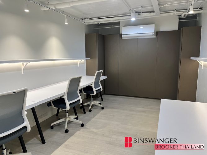 A bright, modern office space with white desks against the wall, grey ergonomic chairs, light wood flooring, and tall built-in storage cabinets. The Binswanger Brooker Thailand logo is in the bottom right corner.