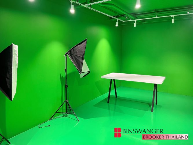 A dedicated green screen studio with bright green walls and flooring, equipped with two professional softbox lighting stands and a minimalist white table in the center. The Binswanger Brooker Thailand logo is in the bottom right corner.