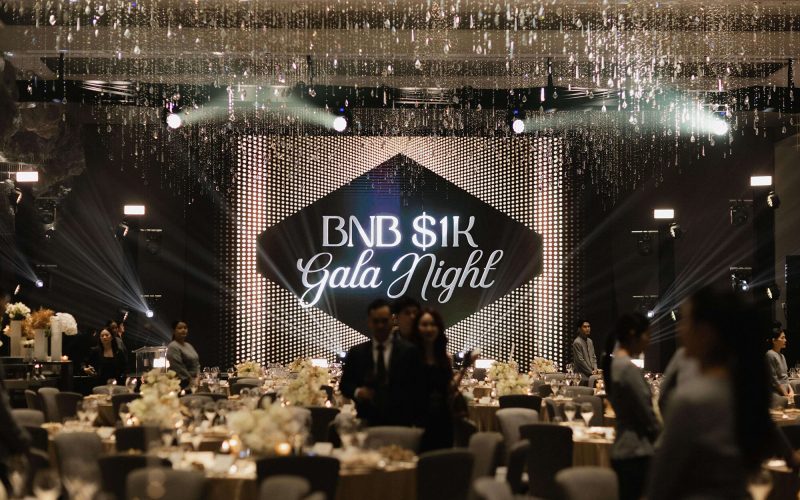 Wide shot of the main ballroom setup for BNB 1K Gala Night hosted by The Brooker Group PCL, featuring a illuminated stage screen and tables set for dinner.