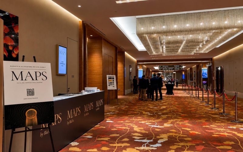 A registration desk and a welcome sign for the Markets & Allocators Private Summit (MAPS) by BluFin, held on July 21, 2026, at Marina Bay Sands, Singapore, set in a grand hotel hallway with a crystal chandelier and patterned carpet.