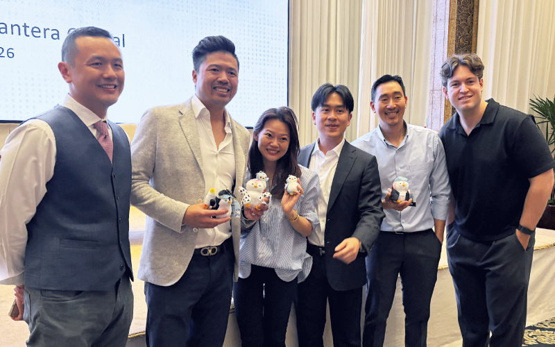 A group of six professionals smiling together at a private business event, with several holding small branded plush figures in front of a presentation screen.